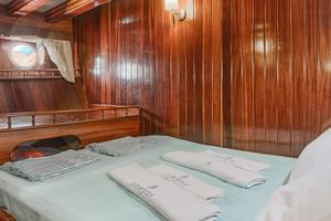 Doppelkabine auf der MS Thalassa mit holzgetäfelten Wänden, Bett mit hellgrüner Bettwäsche, weißen Kissen, Bullaugen und Einbauschränken.