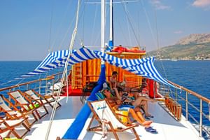 Sonnendeck der MS Mirabela mit Gästen auf Liegestühlen. Blau-weiß gestreifte Markisen spenden Schatten, blaues Meer und Küstenhügel im Hintergrund.