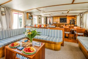 Innenraum des MS Caprice Salons mit blau-weiß gestreiften Polstersitzen, Holzmöbeln, Obsttellern auf dem Couchtisch und Küchenbereich im Hintergrund.