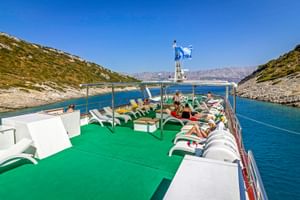 Sonnendeck der MY Mystic mit grünem Boden, weißen Liegen und entspannenden Passagieren. Blaues Küstenwasser und felsige Hügel im Hintergrund.