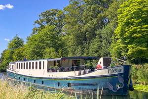 Flusskreuzfahrtschiff MS Fleur an einem von Bäumen gesäumten Kanal in Frankreich. Das weiß-blaue Schiff hat ein offenes Deck mit Passagieren.