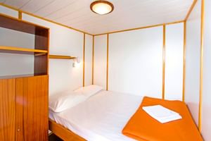 Kompakte Kabine auf der MS Planka mit Doppelbett, weißer Bettwäsche, orangem Handtuch, Holzschrank und weißen Wänden mit Holzverkleidung.