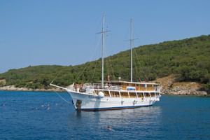 Weißes Motorsegelschiff MS Kazimir mit zwei Masten vor Anker in ruhigem blauen Wasser. Grüne Hügel im Hintergrund, Schwimmer im Wasser.