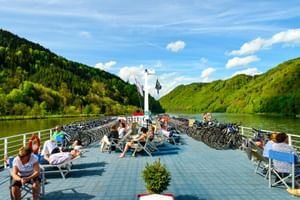 Sonnendeck der MS Normandie mit Passagieren auf Liegestühlen. Fahrräder sind an beiden Seiten geparkt. Grüne Hügel und Fluss im Hintergrund.