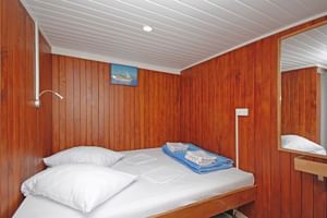 Kompakte Kabine auf der MS Kazimir mit Holzwänden, weißer Decke, Doppelbett mit weißer Bettwäsche, blauen Handtüchern, Leselampe und Spiegel.