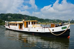 MS Miro, ein weiß-gelbes Flusskreuzfahrtschiff mit schwarzem Rumpf, liegt vor Anker auf ruhigem Wasser mit grünen Hügeln im Hintergrund.