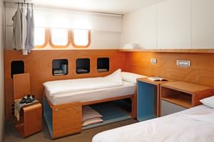 Modern cabin on MS Ave Maria with two beds, wooden walls, storage niches, white cabinets, and a suitcase. Clothes hang by the window.