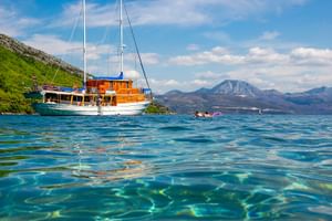 MS Mirabela, ein hölzernes Segelschiff mit weißen Masten, ankert in kristallklarem türkisfarbenem Wasser. Berge und grüne Hügel im Hintergrund.