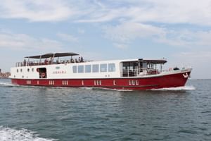 MS Ave Maria Flusskreuzfahrtschiff mit rotem Rumpf und weißem Aufbau auf ruhigem Wasser unter bewölktem Himmel. Passagiere auf Oberdeck sichtbar.
