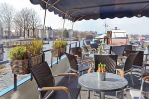 Sonnendeck der MS Fleur mit Korbstühlen und Tischen unter Markise, mit Blick auf Hafen mit Booten und historischen Gebäuden am Wasser.