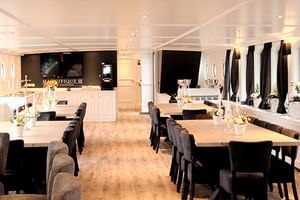 Elegantes Restaurant an Bord der MS Magnifique III mit weißen Tischen, dunklen Stühlen, Bogenfenstern mit schwarzen Vorhängen und Weinregal.