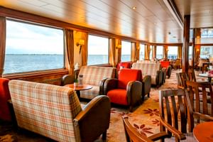 Innenlounge der MS Rigoletto mit karierten Sofas, roten Sesseln und Holzesstischen. Große Panoramafenster bieten Wasserblick.