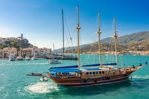 Holzsegelschiff MS Thalassa mit vier Masten in türkisfarbenem Hafenwasser, mit weißer Hangstadt und Bergen im Hintergrund.
