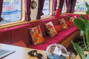 Heller Loungebereich auf der MS Fleur mit rot gepolsterten Sitzbänken, bunten Kissen, großen Fenstern mit bordeauxfarbenen Vorhängen und Grünpflanzen.