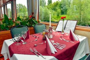 Restaurant an Bord der MS Fluvius mit Tischen, die mit burgunderfarbenen Tischdecken, Weingläsern und Kerzen gedeckt sind. Große Fenster zeigen grüne Ufervegetation.