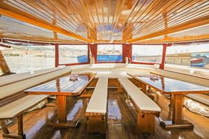 Innenraum des Schiffs MS Sveti Vid mit Holzbänken, Tischen und Lamellendecke. Große Fenster bieten Panoramablick auf den Hafen mit Booten.