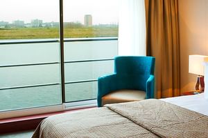 Helle Kabine auf MS SE-Manon mit Doppelbett, großem Panoramafenster mit Flussblick, türkisfarbenem Sessel, Schreibtisch und warmer Beleuchtung.