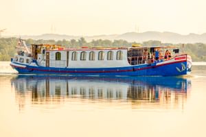 MS L'Estello, ein blau-weißes Flusskreuzfahrtschiff mit Bogenfenstern, gleitet auf ruhigem Wasser mit sichtbarer Spiegelung. Grünes Ufer im Hintergrund.