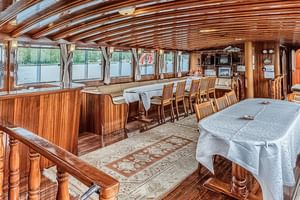 Panoramablick auf den MS Thalassa Salon mit Holzdeckenbalken, großen Fenstern, Esstischen mit weißen Tischdecken und beiger Sitzecke.