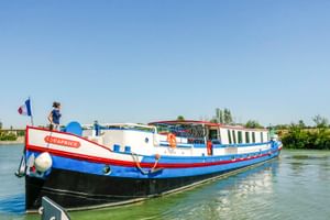 MS Caprice, ein weiß-blaues Flusskreuzfahrtschiff mit rotem Rand, liegt an einem ruhigen Wasserweg. Eine Person steht an Deck unter blauem Himmel.
