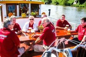 Gruppe von Radfahrern in roten Hemden sitzt an Holztischen auf dem Deck der MS Miro mit Fahrrädern und genießt Getränke an einem sonnigen Tag am Fluss.