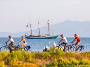 Vier Radfahrer fahren entlang eines Küstenwegs in der Toskana mit einem Dreimaster auf dem Meer und Bergen im Hintergrund.