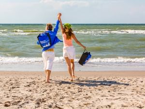 Zwei Personen gehen Hand in Hand am Strand von Nienhagen, halten Taschen und eine blaue Jacke, mit Blick auf die Ostsee.