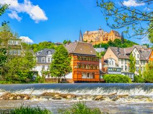 Marburger Landgrafenschloss auf Hügel über bunten historischen Gebäuden entlang der Lahn mit Wehr. Traditionelle Fachwerkhäuser zwischen grünen Bäumen.
