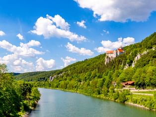 Burg Prunn thront auf weißen Kalksteinfelsen über der Altmühl, umgeben von grünen bewaldeten Hügeln unter blauem Himmel mit weißen Wolken.