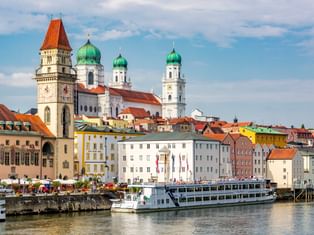 Passauer Altstadt mit bunten Gebäuden an der Donau. Ein Flusskreuzfahrtschiff liegt am Ufer. Kirchtürme mit grünen Kuppeln prägen die Skyline.