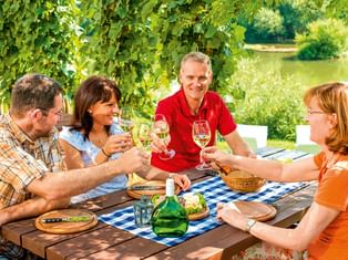 Vier Personen stoßen mit Weißwein an einem Holztisch am Main im Fränkischen Weinland an, mit Weinbergen und Fluss im Hintergrund.