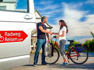 Zwei Frauen geben sich die Hand neben einem Fahrrad und einem Radweg-Reisen.com Transporter. Eine Frau hält Gepäck, die andere steht bei einem Tourenrad.