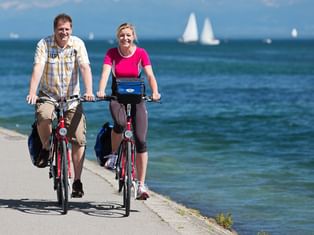 Zwei Radfahrer fahren auf einem gepflasterten Uferweg am Bodensee mit Segelbooten auf blauem Wasser und klarem Himmel im Hintergrund.