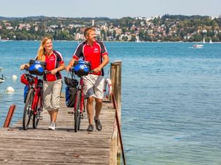 Zwei Radfahrer in roten Trikots gehen mit Rädern auf Holzsteg am Bodensee in Dingelsdorf. See, Boote und Hangstadt im Hintergrund sichtbar.