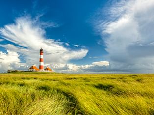 Leuchtturm Westerhever mit rot-weißen Streifen und Nebengebäuden in grüner Salzwiese unter dramatischem Wolkenhimmel an der Nordsee.