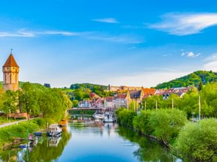 Malerischer Blick auf Wertheim mit dem Main, Booten am grünen Ufer, einem mittelalterlichen Steinturm und historischen Gebäuden unter blauem Himmel.