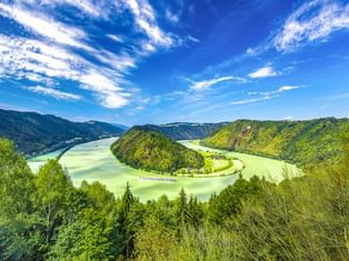 Luftaufnahme der Schlögener Schlinge, einer dramatischen Hufeisenschleife der Donau, umgeben von bewaldeten Hügeln unter blauem Himmel.