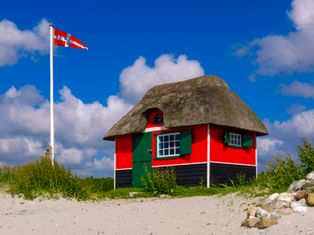 Rot-schwarze Strandhütte mit Reetdach und grünen Fensterläden am Sandstrand. Dänische Flagge weht an weißem Mast unter blauem Himmel mit Wolken.