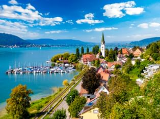 Luftaufnahme von Sipplingen am Bodensee mit weißem Kirchturm, Jachthafen mit Segelbooten und Bergen im Hintergrund unter blauem Himmel.