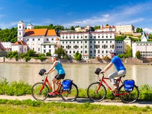 Zwei Radfahrer mit roten Rädern und blauen Taschen fahren am Inn in Passau entlang, mit historischen Gebäuden und einer Kirche am gegenüberliegenden Ufer.