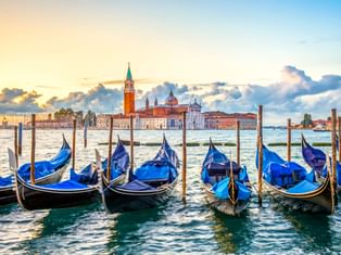 Blaue Gondeln an Holzpfählen in Venedig mit der Kirche San Giorgio Maggiore über der Lagune unter dramatischem Abendhimmel.