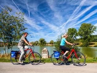 Zwei Radfahrer auf roten Rädern mit blauen Taschen fahren an einem See nahe der Altmühl-Quelle. Ein Steinmarker steht zwischen ihnen unter blauem Himmel.