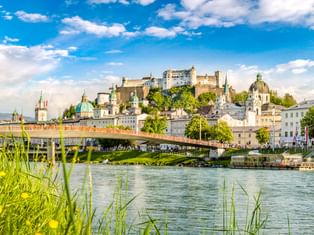 Blick über die Salzach auf Salzburgs Altstadt mit Festung Hohensalzburg auf dem Hügel, historischen Kirchen und Brücke, eingerahmt von grünem Ufer.