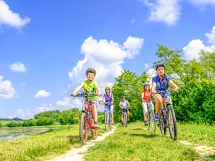 Five cyclists with helmets riding on a grass path beside a river under blue sky with white clouds. Green trees and hills in background.
