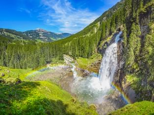 Krimmler Wasserfälle stürzen über Felsklippe mit Regenbogen im Sprühnebel, umgeben von grünen Wiesen und dichten Wäldern in den Alpen.