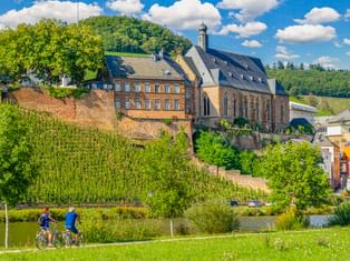 Zwei Radfahrer auf Weg entlang der Saar mit St. Laurentius Kirche am Hang in Saarburg, umgeben von Weinbergen und grünen Hügeln unter blauem Himmel.