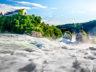 Rheinfall in Schaffhausen mit tosenden Wassermassen, Felsinseln und historischem Schloss auf bewaldetem Hügel unter blauem Himmel.