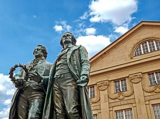 Bronzestatue von Goethe und Schiller, die nebeneinander vor dem Nationaltheater in Weimar unter blauem Himmel mit weißen Wolken stehen.