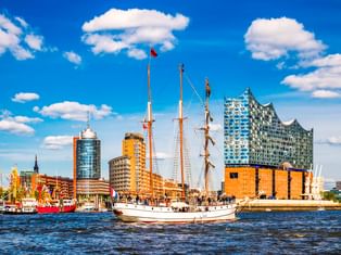 Weißes Segelschiff auf der Elbe mit Hamburgs Elbphilharmonie und historischen Hafengebäuden unter blauem Himmel mit Wolken.