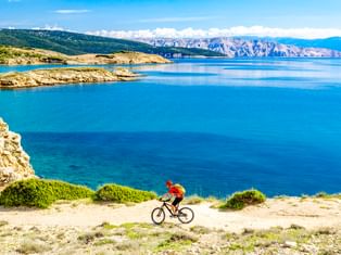 Radfahrer in roter Jacke fährt entlang Küstenweg mit Blick auf türkisfarbene Adria mit Felsklippen und Bergen in Kroatien.